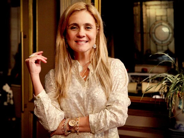 Personagem Carminha com camisa clara e joias luxuosas em ambiente sofisticado da novela Avenida Brasil.