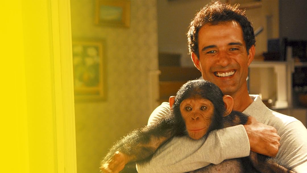 Homem sorridente segurando um chimpanzé dentro de casa na novela Caras & Bocas